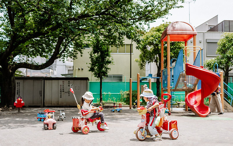 Two toddlers ride tricycles in a colorful outdoor playground surrounded by trees and play equipment. A bright red slide and climbing structure are visible in the background, adding to the cheerful atmosphere of the park.
